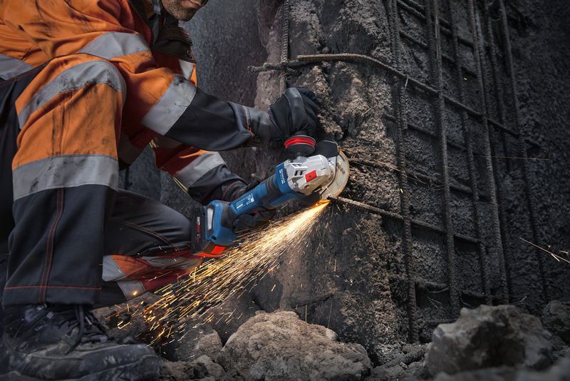 GWS18V-11PS A person wearing safety equipment uses an angle grinder to cut concrete and exposed rebar.