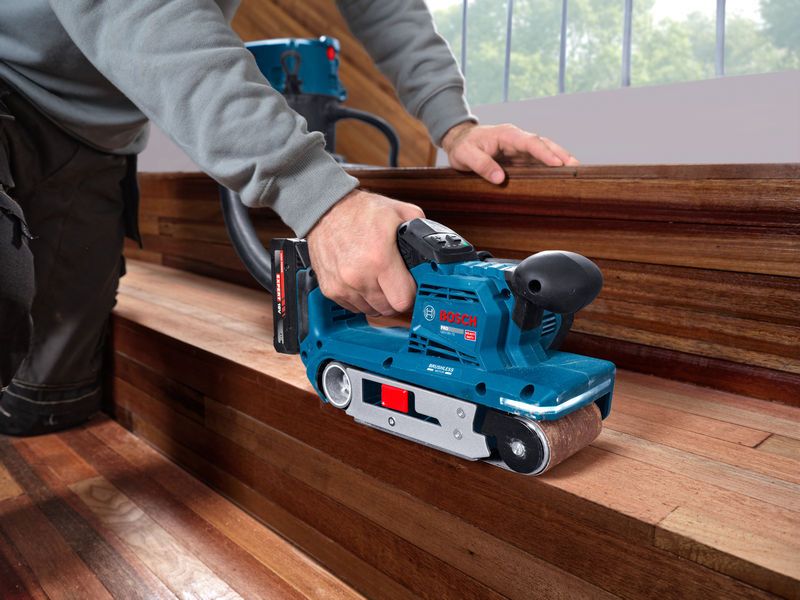 A person uses a belt sander to smooth wooden stairs indoors.