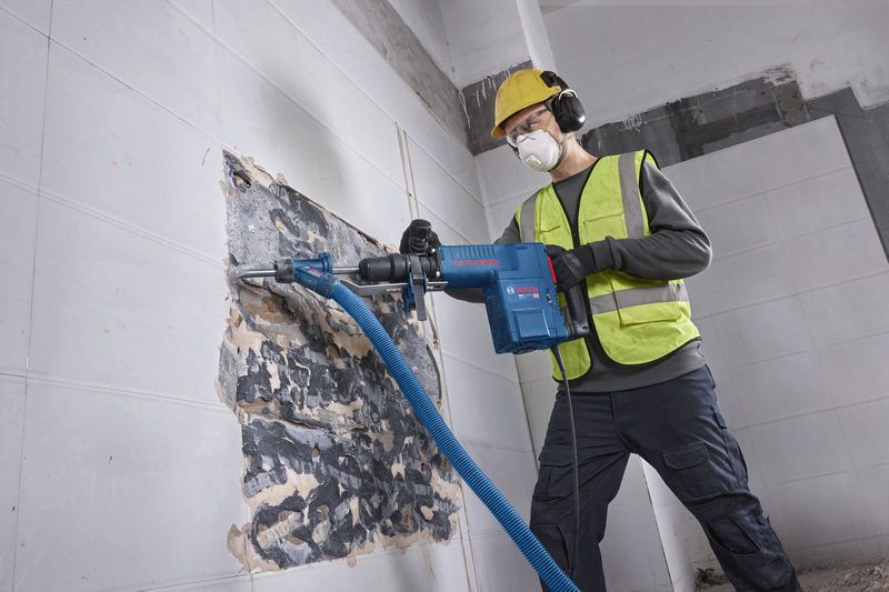 A person wearing safety equipment uses a demolition hammer to break wall tiles.