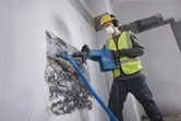 A person wearing safety equipment uses a demolition hammer to break wall tiles.