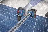 A person wearing safety equipment uses a cordless drill on a solar panel frame.