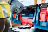 A person wearing safety equipment lifts a blue and red carrying case system.