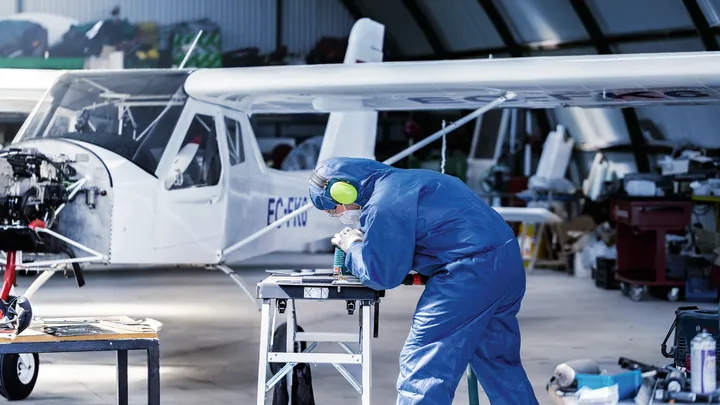 Person wearing safety equipment drills a metal part on a workbench near a small aircraft.