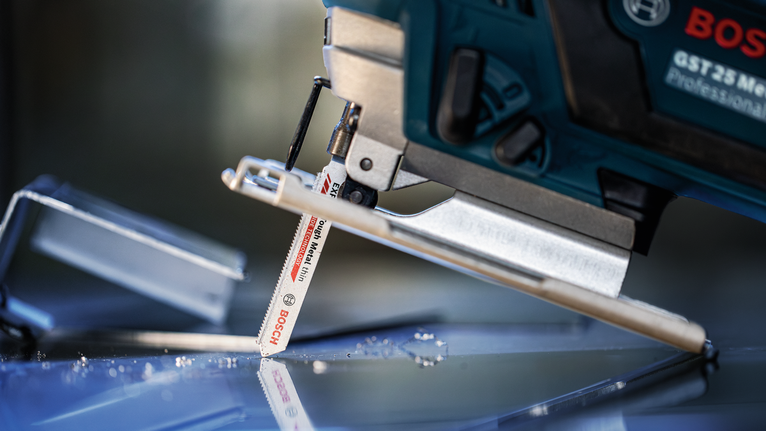 A jigsaw cuts through a sheet of glass with scattered glass shards nearby.
