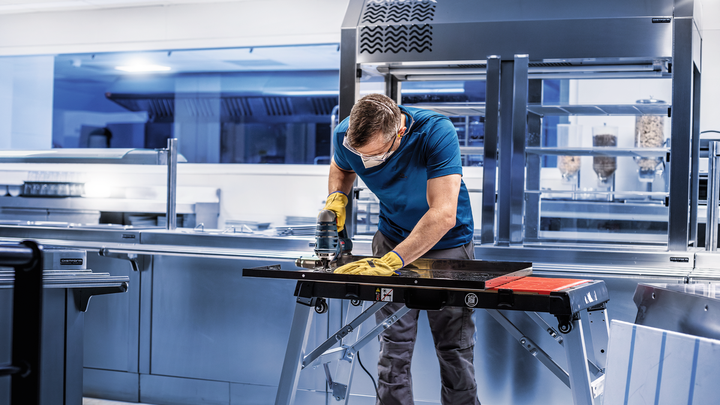 A person wearing safety equipment grinds a metal sheet on a workbench in a kitchen.