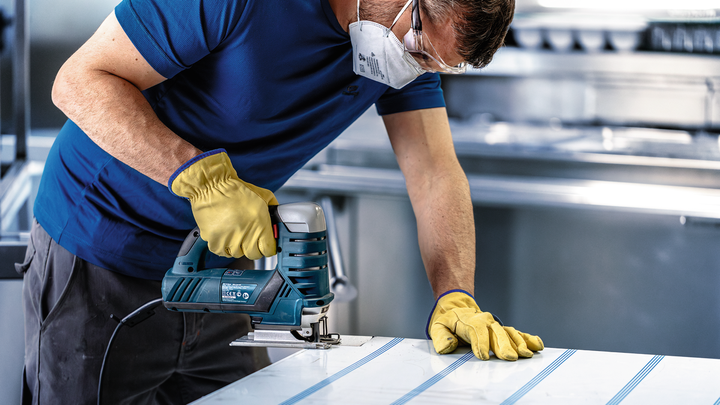 Person wearing safety equipment uses a jigsaw to cut sheet material.