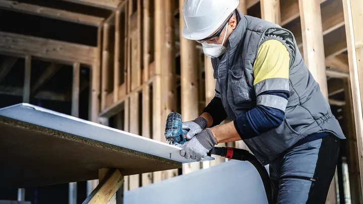 Object #200597853: 2793289_app_web_06 Person wearing safety equipment uses a power drill on a plasterboard in a building frame.