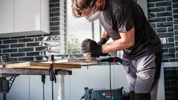 Object #200597888: 2793295_app_web_05 Person wearing safety equipment uses a jigsaw to cut wood on a workbench.