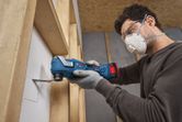 A person wearing safety equipment cuts a square opening in drywall with a multifunction tool.