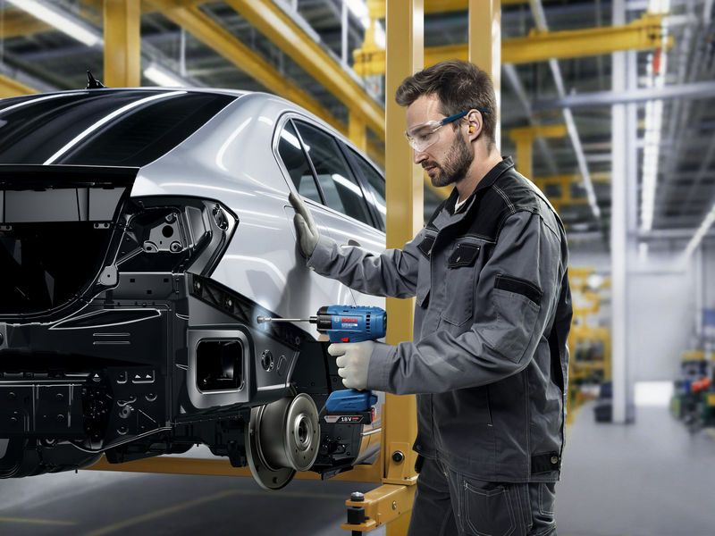 A person wearing safety equipment uses a cordless impact wrench on a car frame.