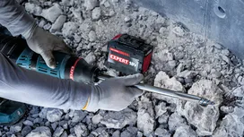 Person wearing safety equipment operates a cordless rotary hammer on rocky ground.