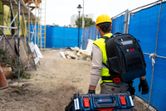 A person wearing safety equipment carries a contractor backpack and carrying case system at a construction site.