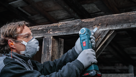 Person wearing safety equipment uses a cordless tool to sand weathered wood.