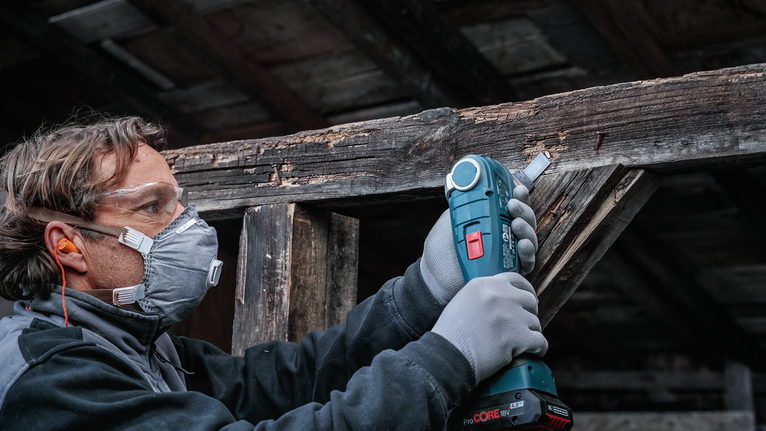 Person wearing safety equipment uses a cordless tool to sand weathered wood.