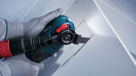 Person wearing safety equipment cuts a square opening in drywall using a power tool.