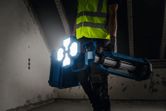 A person wearing safety equipment carries a cordless jobsite light in a dimly lit room.