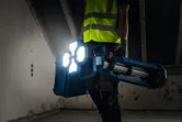 A person wearing safety equipment carries a cordless jobsite light in a dimly lit room.