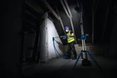 A person wearing safety equipment drills into a wall, illuminated by a cordless jobsite light.