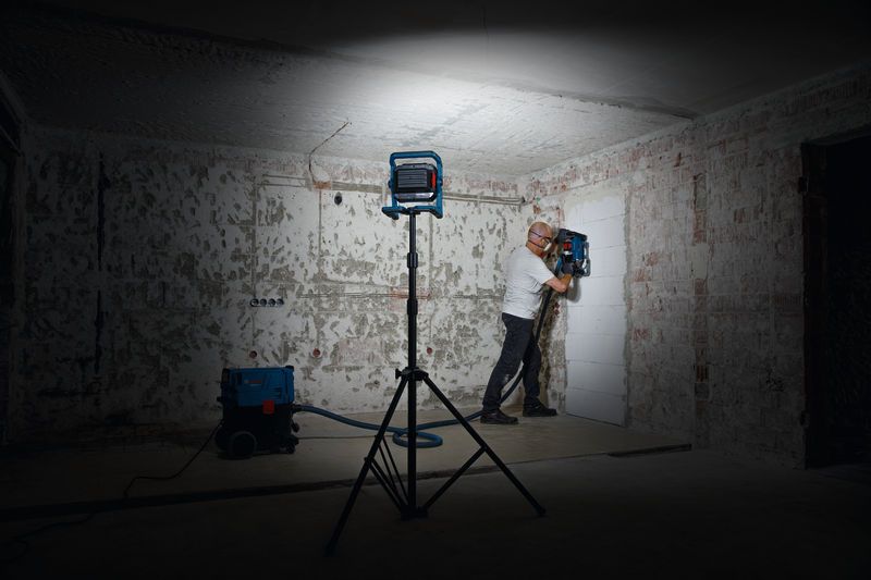 A person wearing safety equipment operates a wall drilling tool under work light.