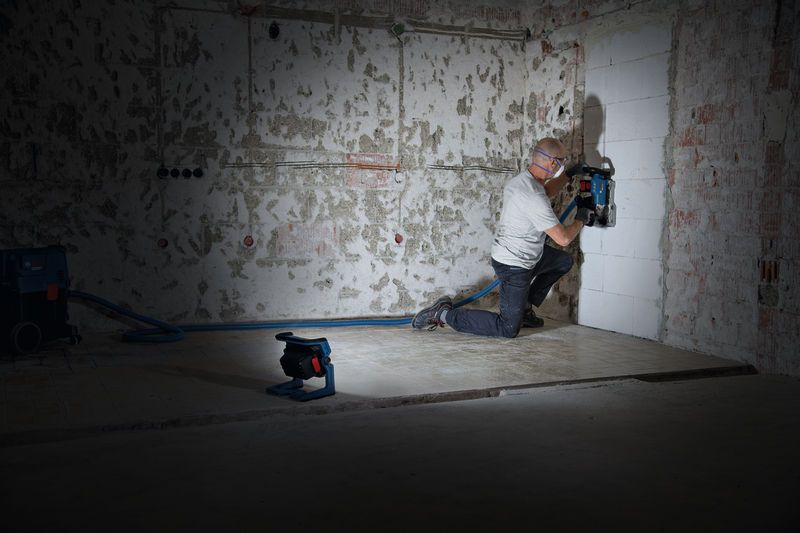 A person wearing safety equipment cuts channels into a wall, illuminated by a cordless jobsite light.