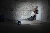 A person wearing safety equipment cuts channels into a wall, illuminated by a cordless jobsite light.