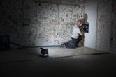 A person wearing safety equipment cuts channels into a wall, illuminated by a cordless jobsite light.