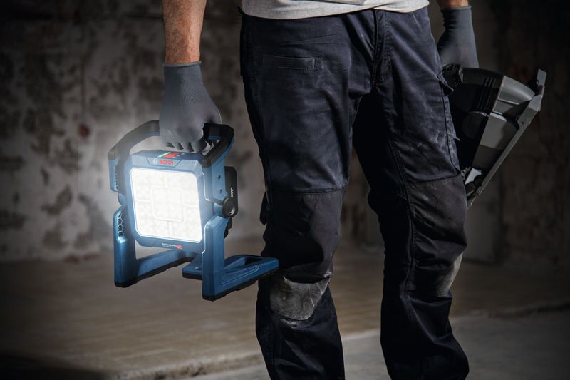 A person wearing safety equipment holds a cordless jobsite light and a power tool.