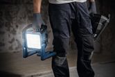 A person wearing safety equipment holds a cordless jobsite light and a power tool.