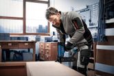 A person wearing safety equipment uses a cordless brad nailer on wooden furniture.