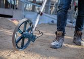 A person in work boots uses a measuring wheel on a sandy construction site.