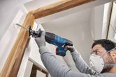 A person wearing safety equipment uses a cordless reciprocating saw to cut a door frame.