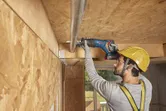 A person wearing safety equipment uses a cordless reciprocating saw to cut a ceiling pipe.