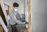 A person wearing safety equipment uses a cordless reciprocating saw to cut a wooden frame.
