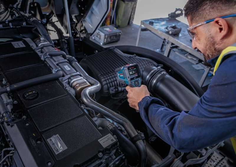 A person wearing safety equipment uses a thermo detector to check engine temperature.