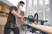 A person wearing safety equipment cuts wood with a mitre saw in a bright workshop.