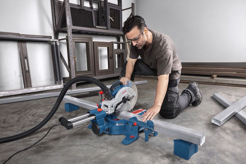 A person wearing safety equipment uses a mitre saw to cut a metal beam in a workshop.