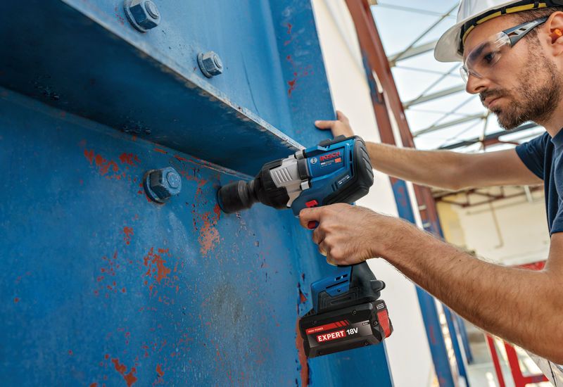 A person wearing safety equipment uses an impact wrench to fasten bolts on a large metal beam.
