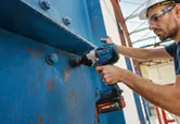 A person wearing safety equipment uses an impact wrench to fasten bolts on a large metal beam.