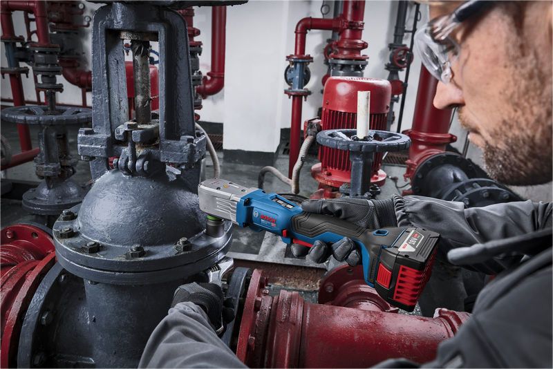 A person wearing safety equipment uses a cordless impact wrench on industrial piping.