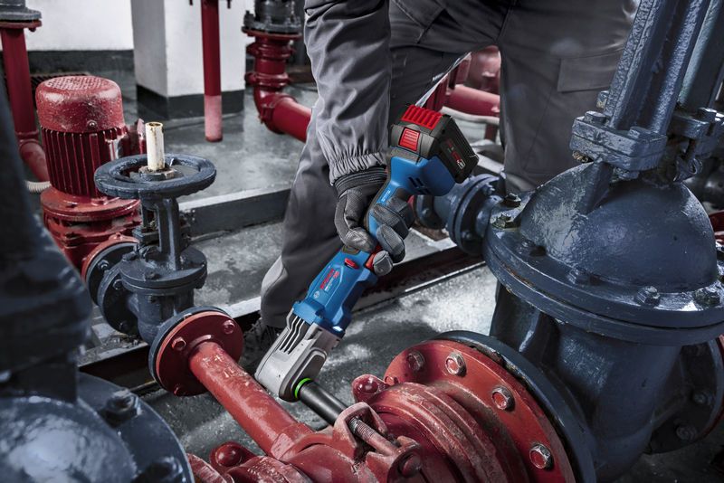 A person wearing safety equipment uses a cordless impact wrench on industrial piping.