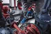 A person wearing safety equipment uses a cordless impact wrench on industrial piping.