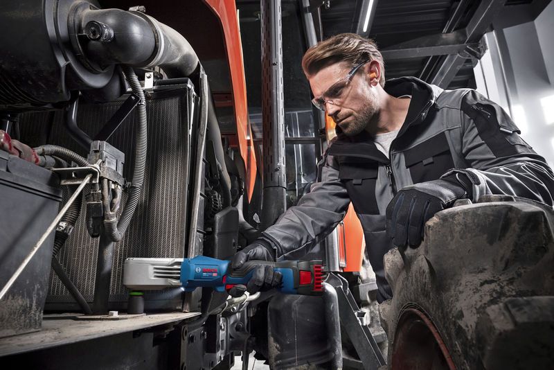 A person wearing safety equipment uses a cordless impact wrench to fasten parts on heavy machinery.