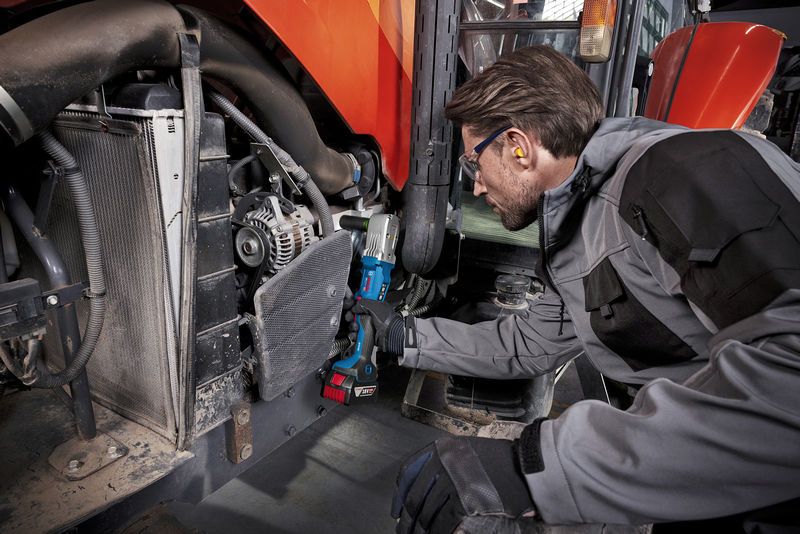 A person wearing safety equipment uses a cordless impact wrench on heavy machinery.