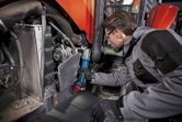 A person wearing safety equipment uses a cordless impact wrench on heavy machinery.