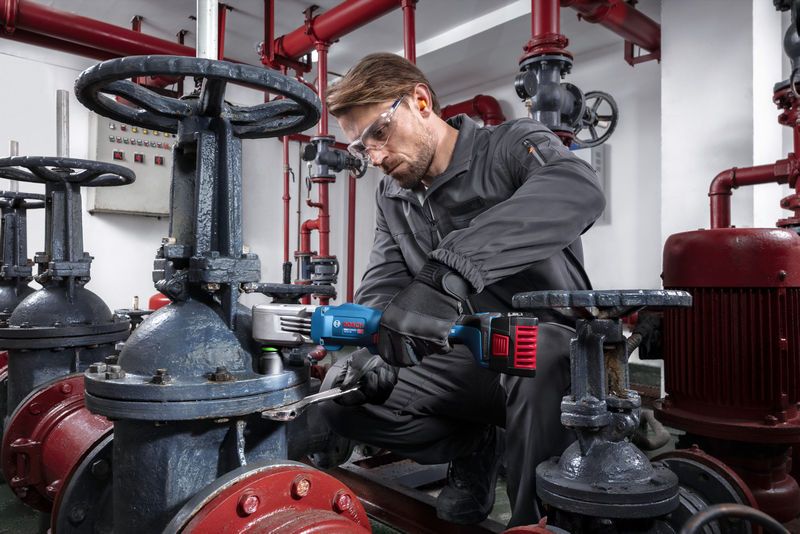 A person wearing safety equipment uses a cordless impact wrench to tighten a valve in an industrial setting.