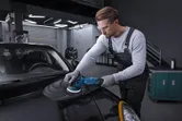 A person wearing safety equipment polishes a black car hood with a power polisher.