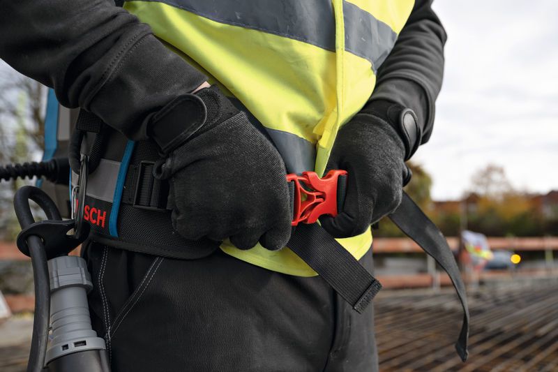 A person wearing safety equipment adjusts a belt with an attached backpack on a construction site.