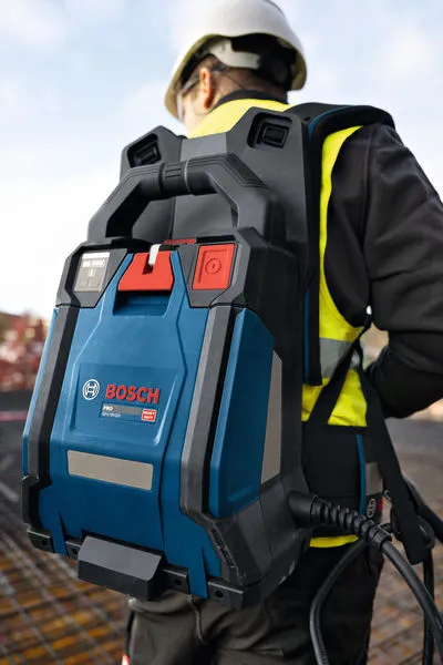 A person wearing safety equipment carries a blue backpack concrete vibrator at a construction site.