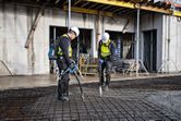 Two workers wearing safety equipment use power tools to consolidate concrete on a rebar slab.