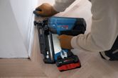 A person wearing safety equipment uses a cordless finish nailer on floor trim.